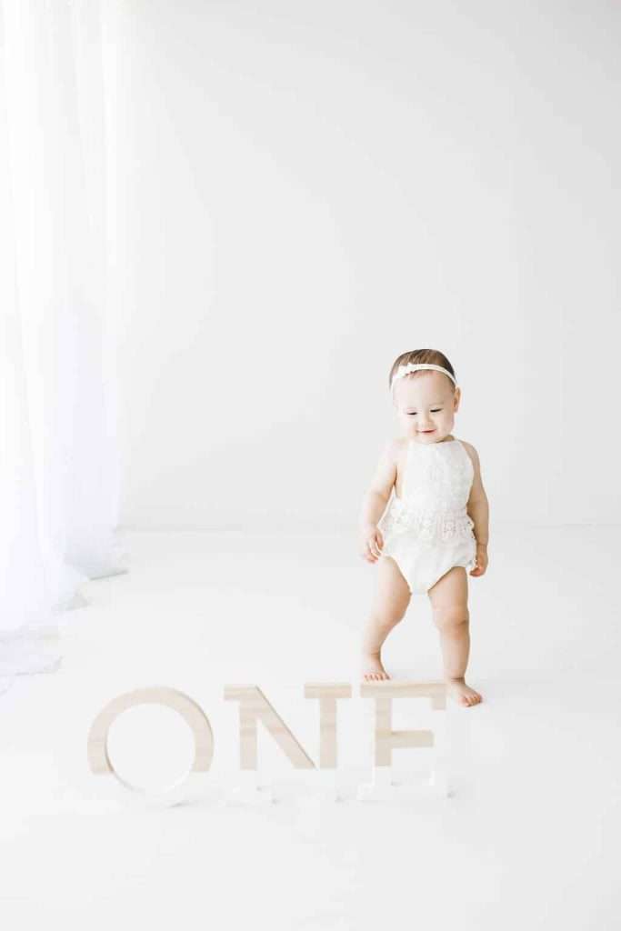 A baby in a white outfit stands next to large wooden letters spelling “ONE” in a bright, minimalistic room.