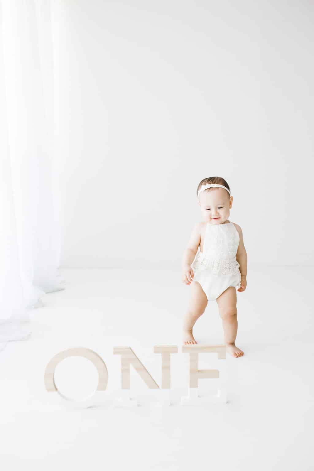 A baby in a white outfit stands next to large wooden letters spelling “ONE” in a bright, minimalistic room.