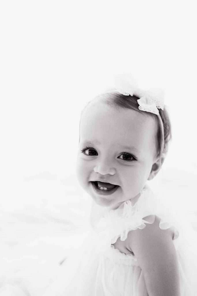 Smiling baby wearing a white dress and a headband with a bow, photographed in black and white against a light background.