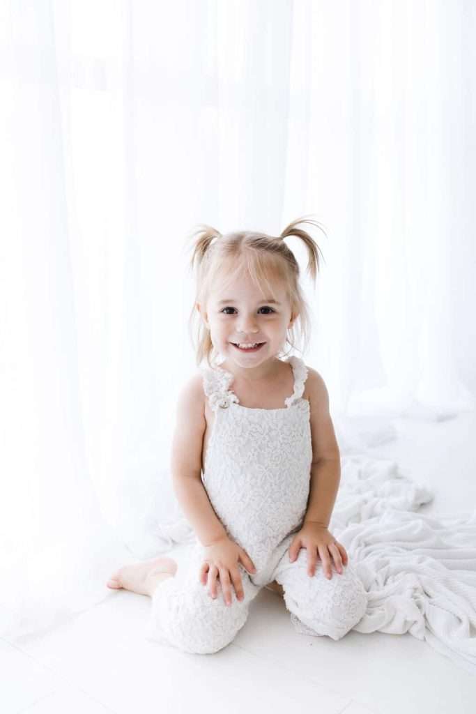 Young child with blonde hair in pigtails, wearing a white textured outfit, sits smiling on a white blanket in a bright, softly lit room.