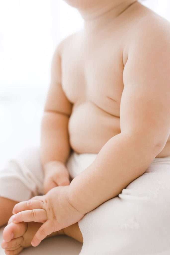 A close-up of a baby sitting, showing the baby’s chubby arms, legs, and torso. The baby is wearing light-colored pants. The background is softly blurred.