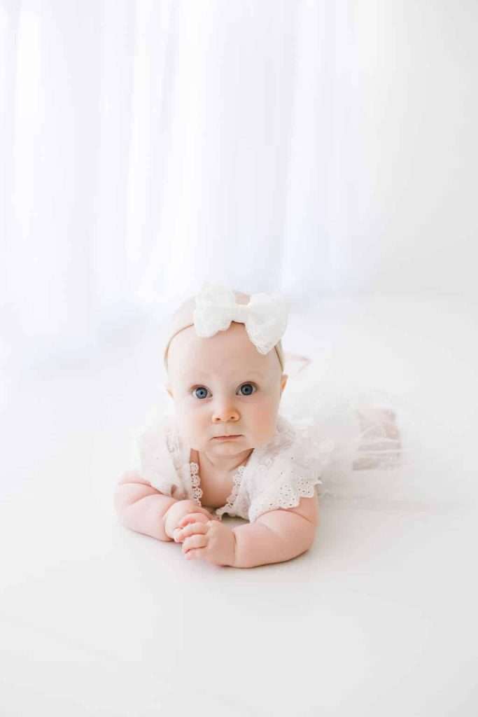 A baby wearing a white dress and a white bow headband lies on their stomach against a white background.
