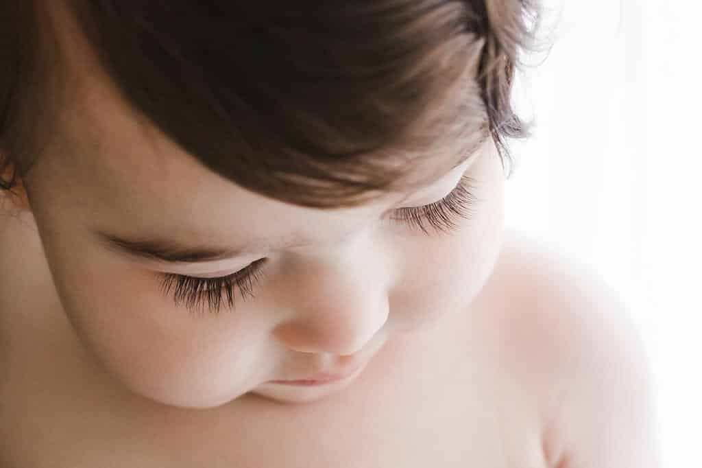 Close-up of a young child’s face looking down, showing long eyelashes, soft skin, and brown hair against a white background.