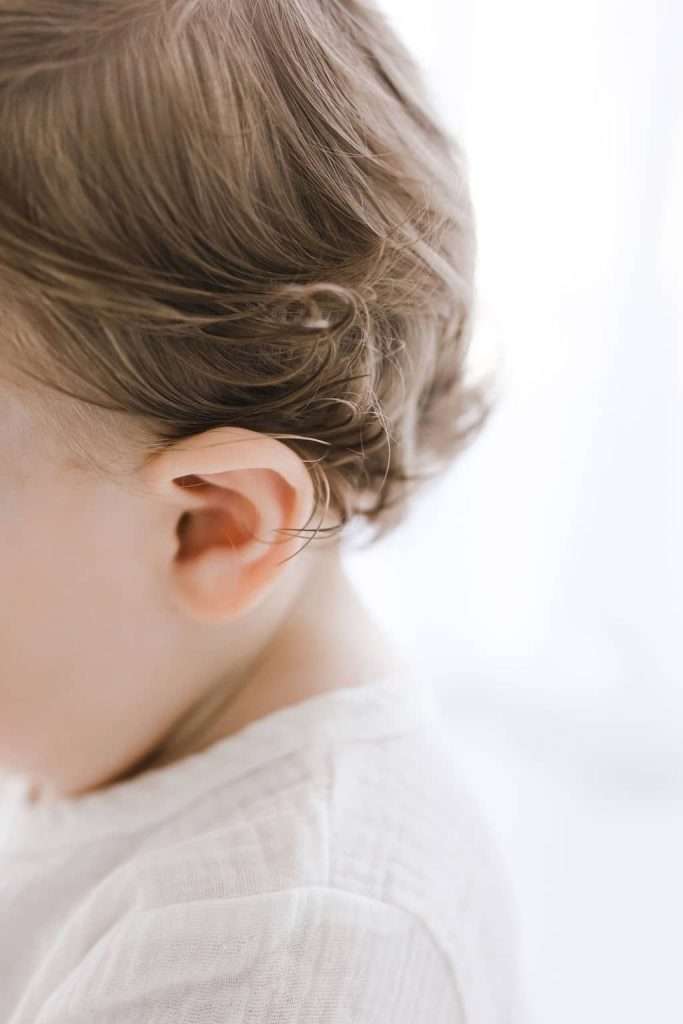 Close-up of a young child’s head and ear, showing light brown wavy hair and a white shirt against a bright, soft-focus background.