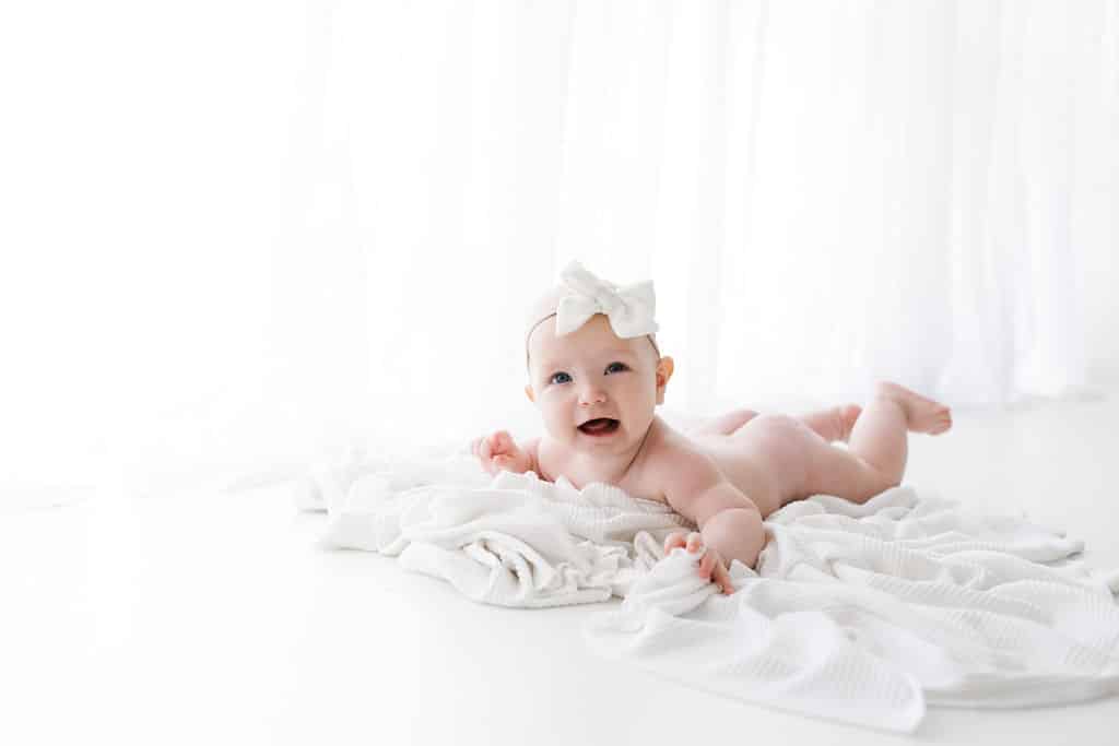 A baby with a white bow headband lies on a white blanket, smiling, in a bright, white room.