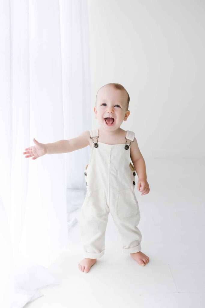 Smiling baby wearing white overalls stands barefoot on a white floor, holding one arm out next to a sheer white curtain.