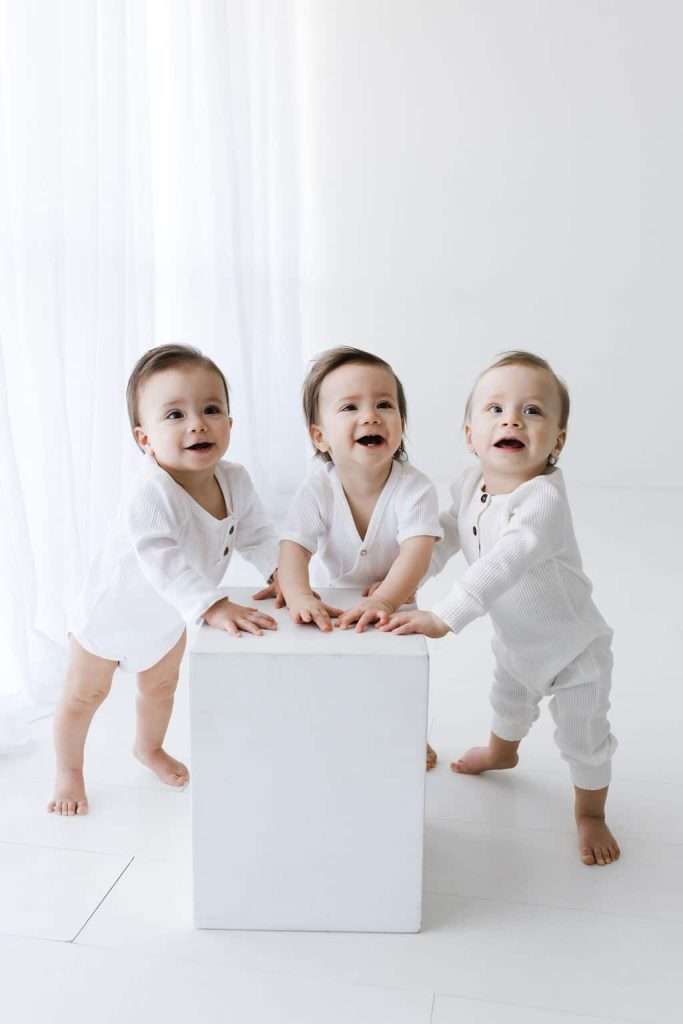 Three young children in white outfits stand around a white cube in a bright, minimalist room with white curtains and floor.