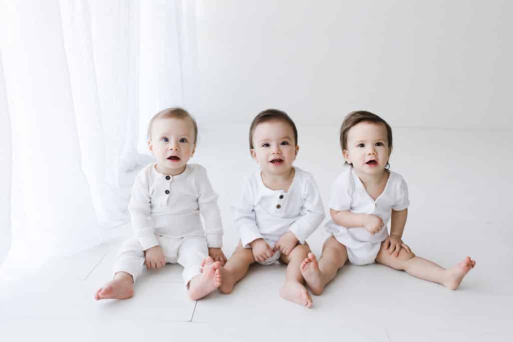 Three babies dressed in white outfits sit barefoot on a white floor against a white background, looking toward the camera.