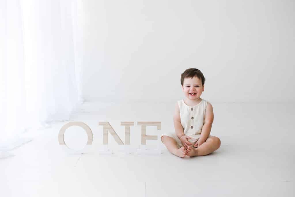 A smiling toddler sits barefoot on the floor in front of large letters spelling “ONE” against a white background.