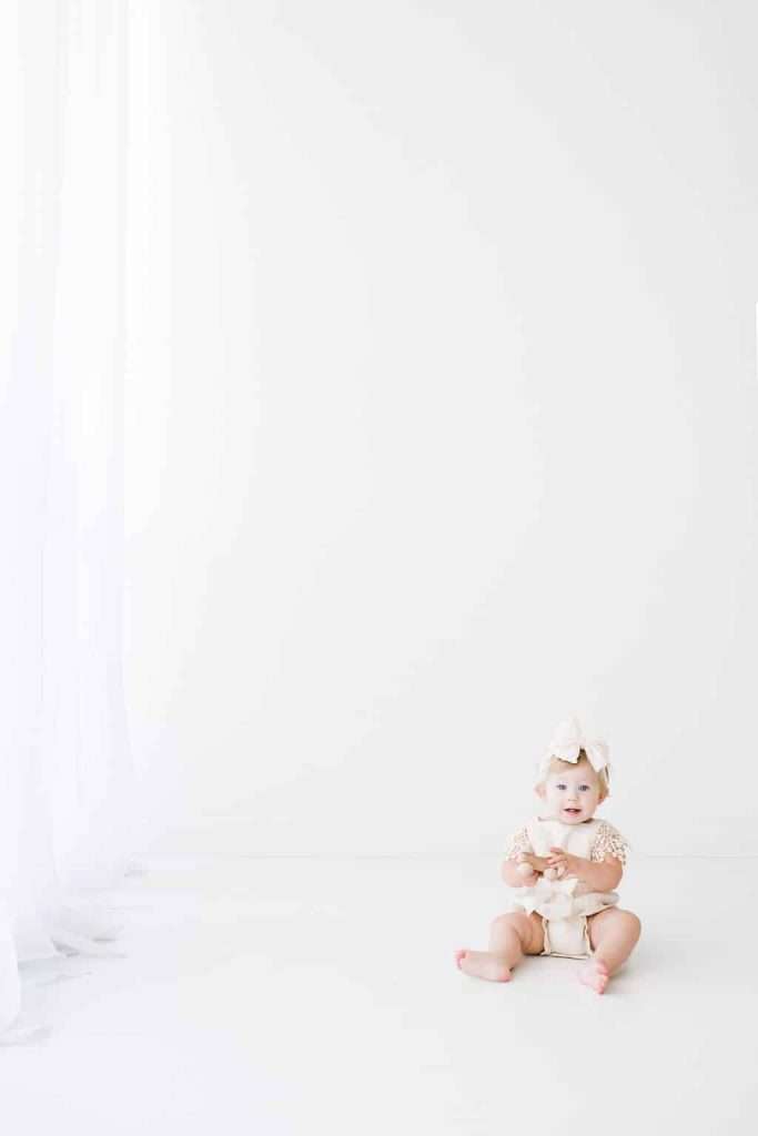 A baby wearing a bow headband and neutral clothing sits on the floor against a plain white background, holding a small object.