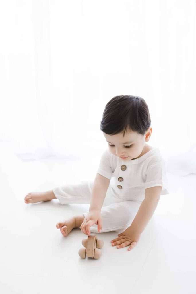 A young child in a white outfit sits on the floor playing with a small wooden toy car against a bright, white background.