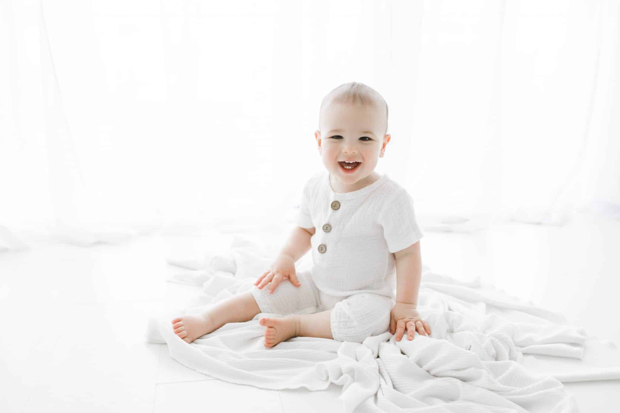A baby in a white outfit smiles while sitting on a white blanket in a bright, softly lit room.