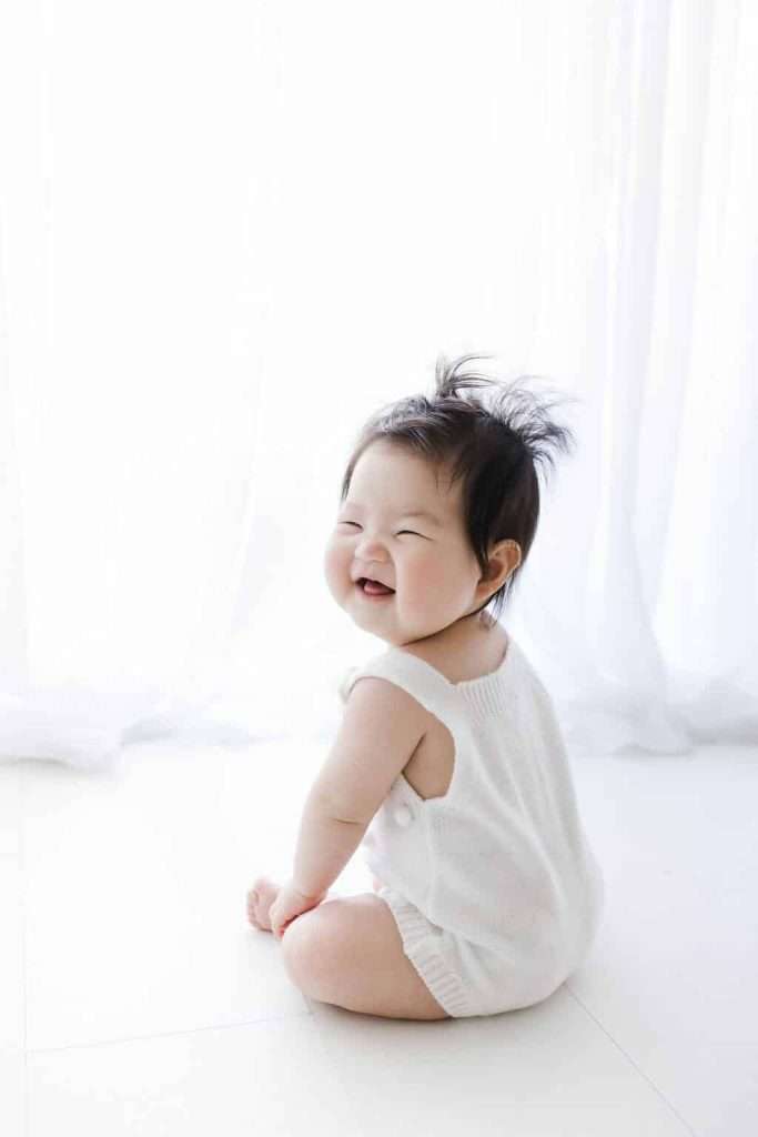 A baby wearing a white romper sits on the floor, smiling and looking over their shoulder in front of a bright white curtain.