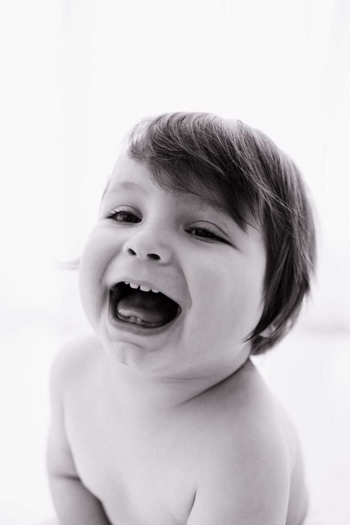 A laughing baby with short, tousled hair looks slightly upward in a close-up, black-and-white photograph against a light background.