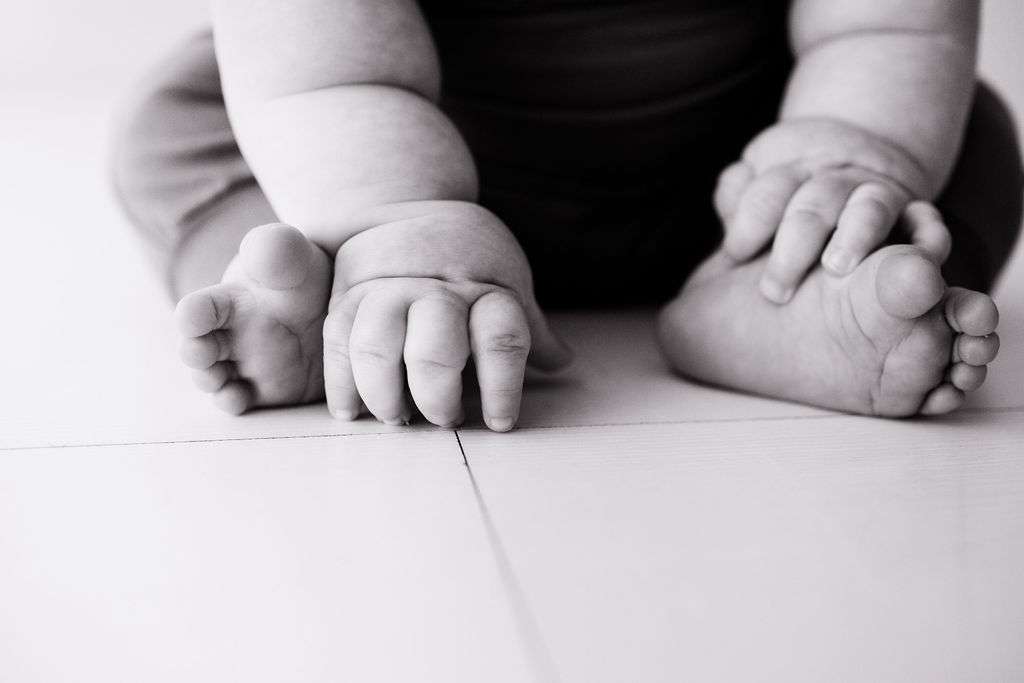 Close-up black and white photo of a baby sitting on the floor, showing chubby hands touching the ground and bare feet crossed.