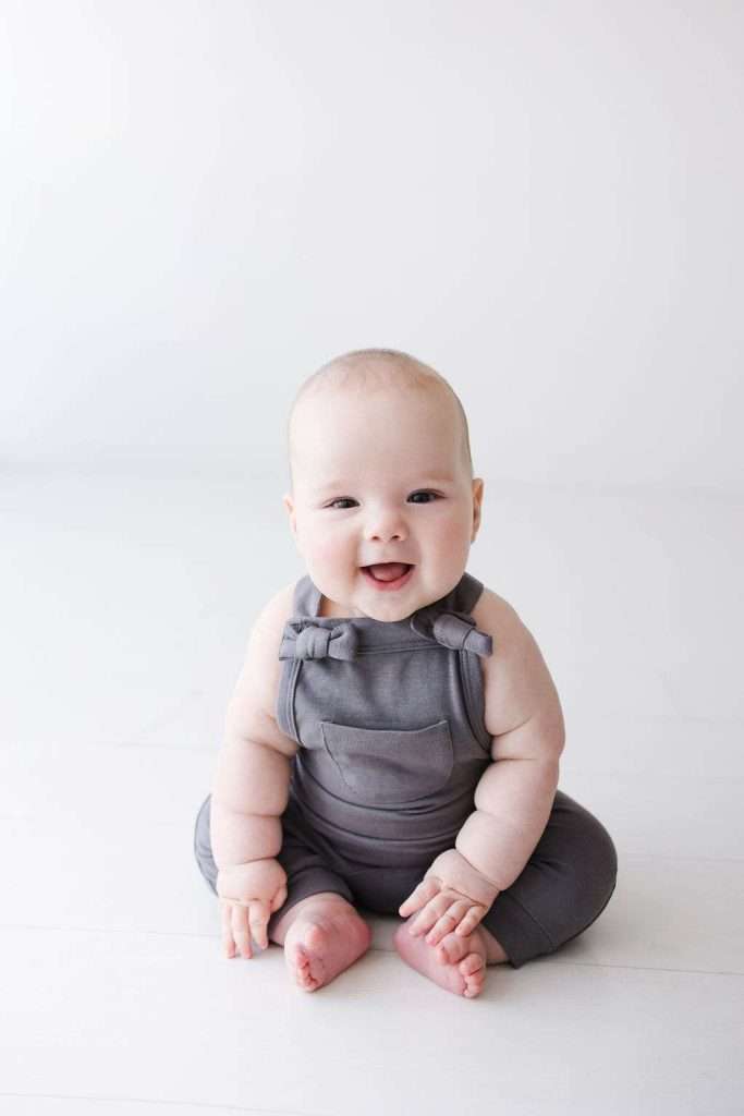 A baby wearing gray overalls sits on a white floor, smiling and looking forward against a plain light background.