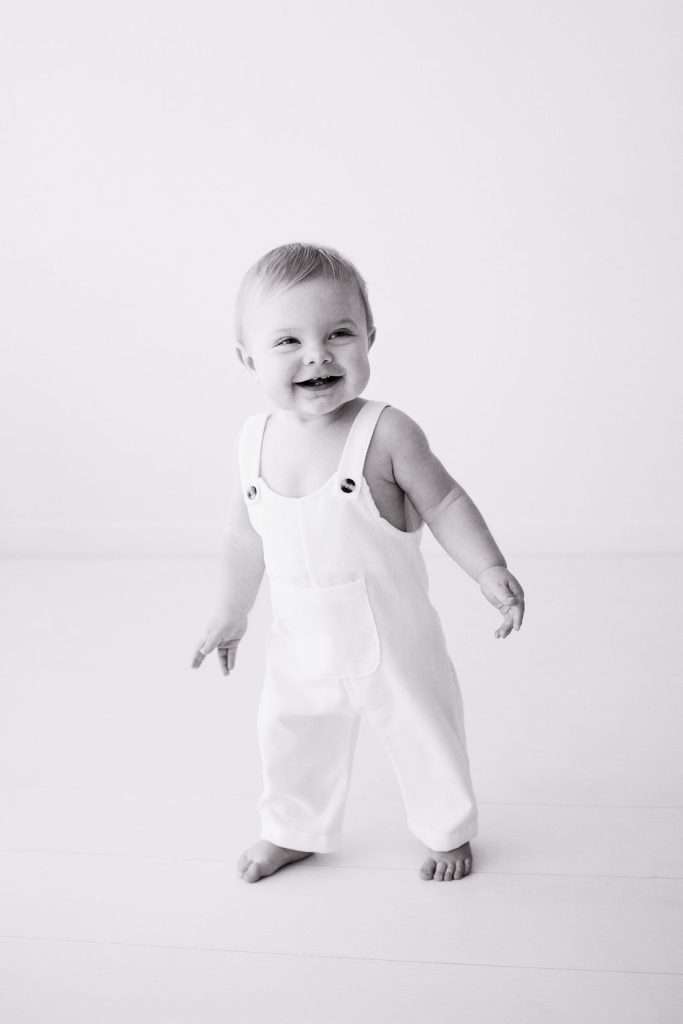 Smiling baby standing barefoot in white overalls against a plain white background, looking slightly to the side.