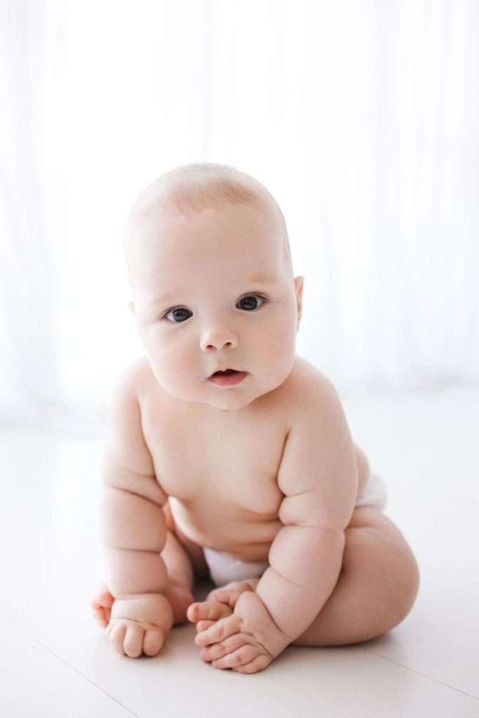 A baby with light skin and no hair wearing a diaper sits on a white surface, looking toward the camera with a neutral expression. A white curtain background is softly lit.