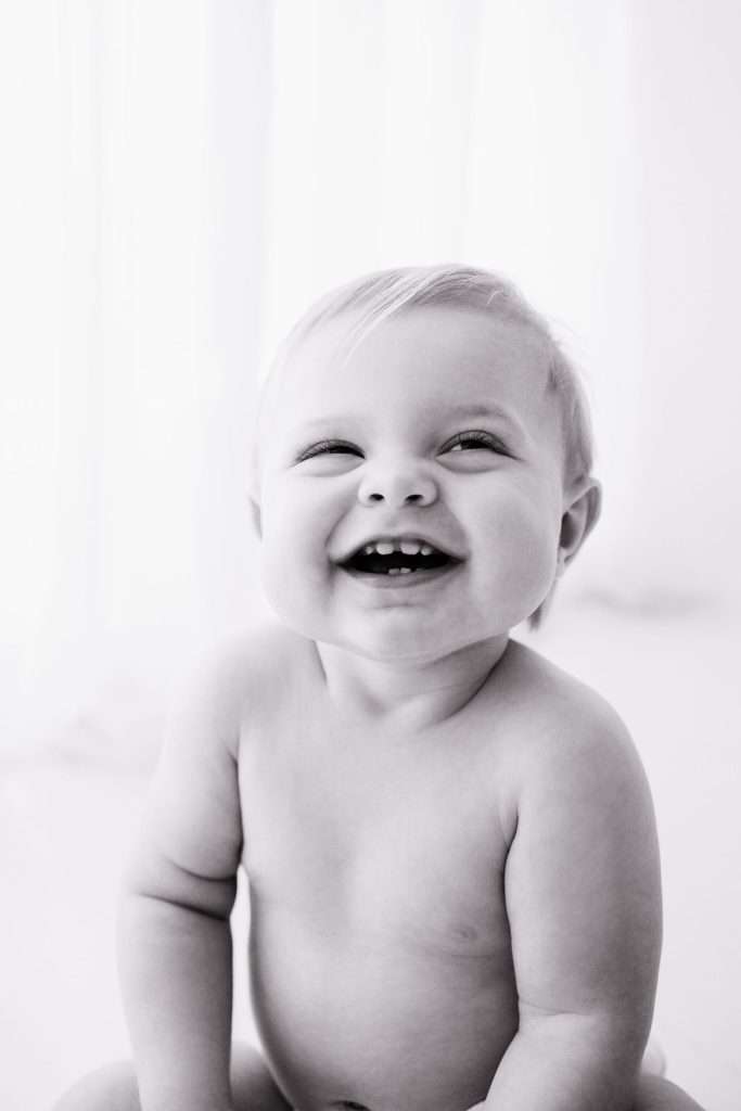Black and white photo of a smiling baby sitting upright, looking slightly upward, with visible teeth and short, light hair.