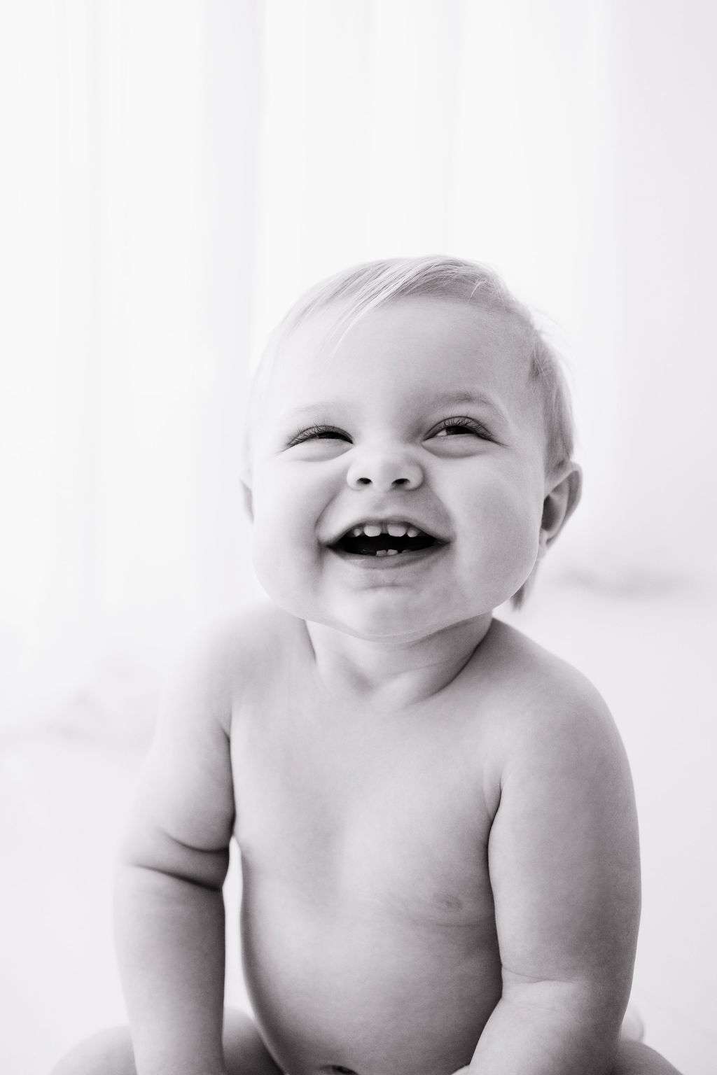 Black and white photo of a smiling baby sitting upright, looking slightly upward, with visible teeth and short, light hair.