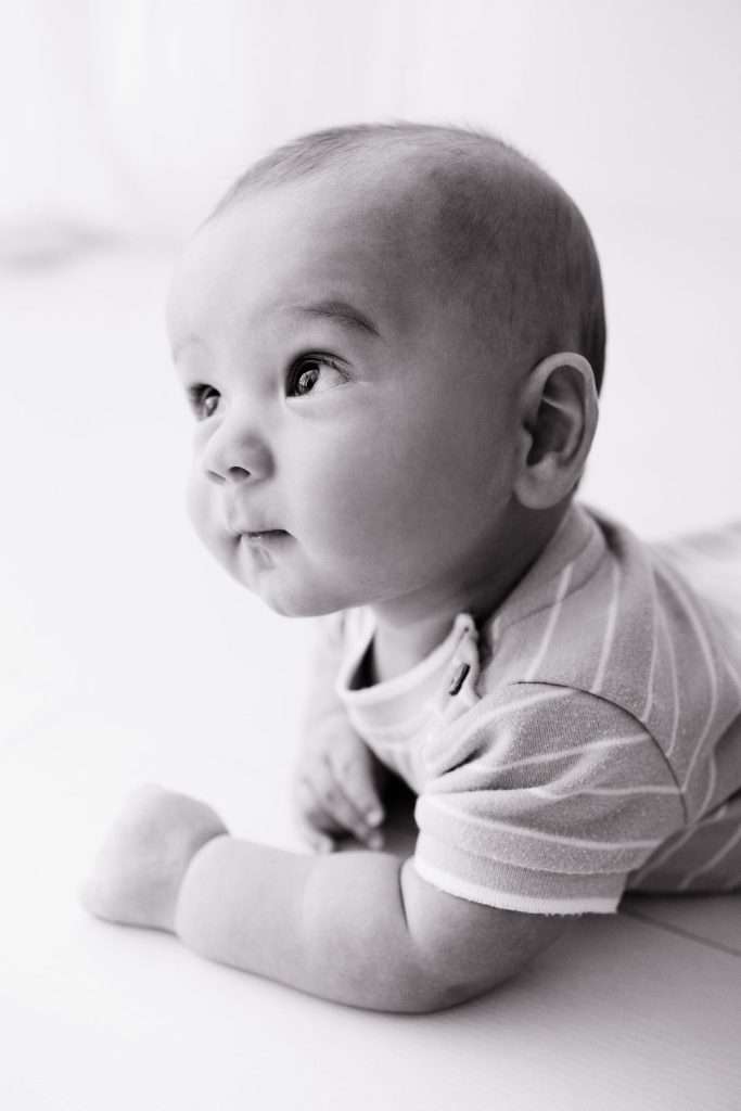 Black and white photo of a baby lying on their stomach, wearing a striped shirt, looking up and to the side with a curious expression.