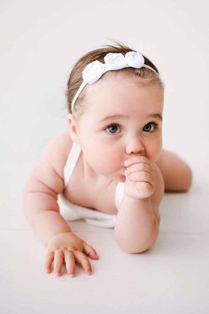 A baby wearing a white headband with three flowers lies on their stomach, looking up and sucking their thumb.