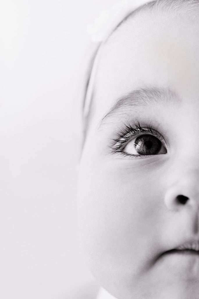 Close-up, black-and-white photo of a baby’s face, showing one eye, eyebrow, part of the nose, and lips. The background is light and out of focus.