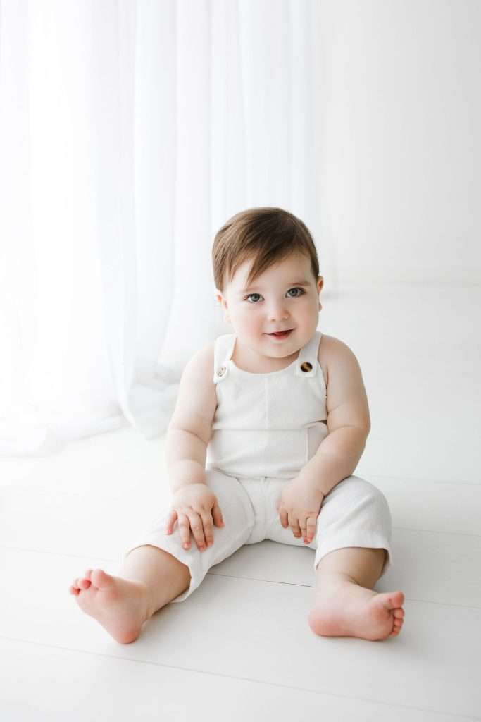 A baby with brown hair, dressed in white overalls, sits on a white floor in a bright room with sheer curtains.