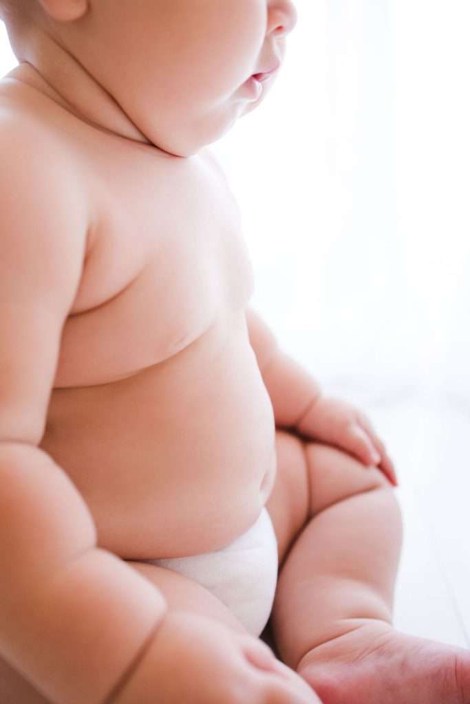 Close-up of a baby sitting while wearing a diaper, showing the baby’s chubby arms, torso, and legs in natural light.