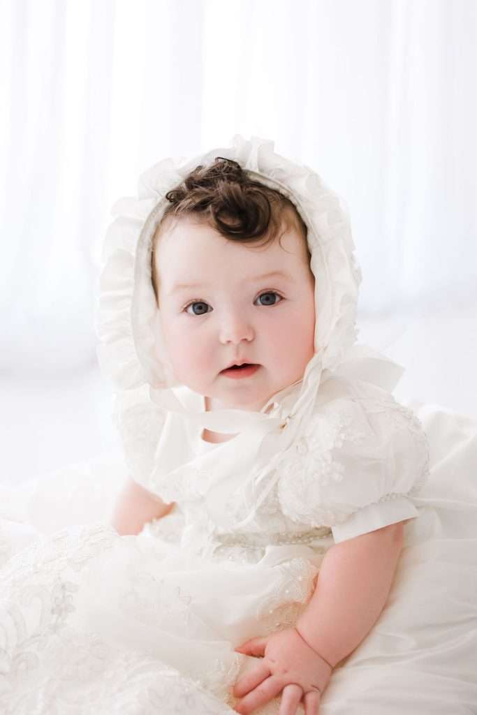 Baby wearing a white bonnet and lace dress sits against a light background, looking toward the camera.