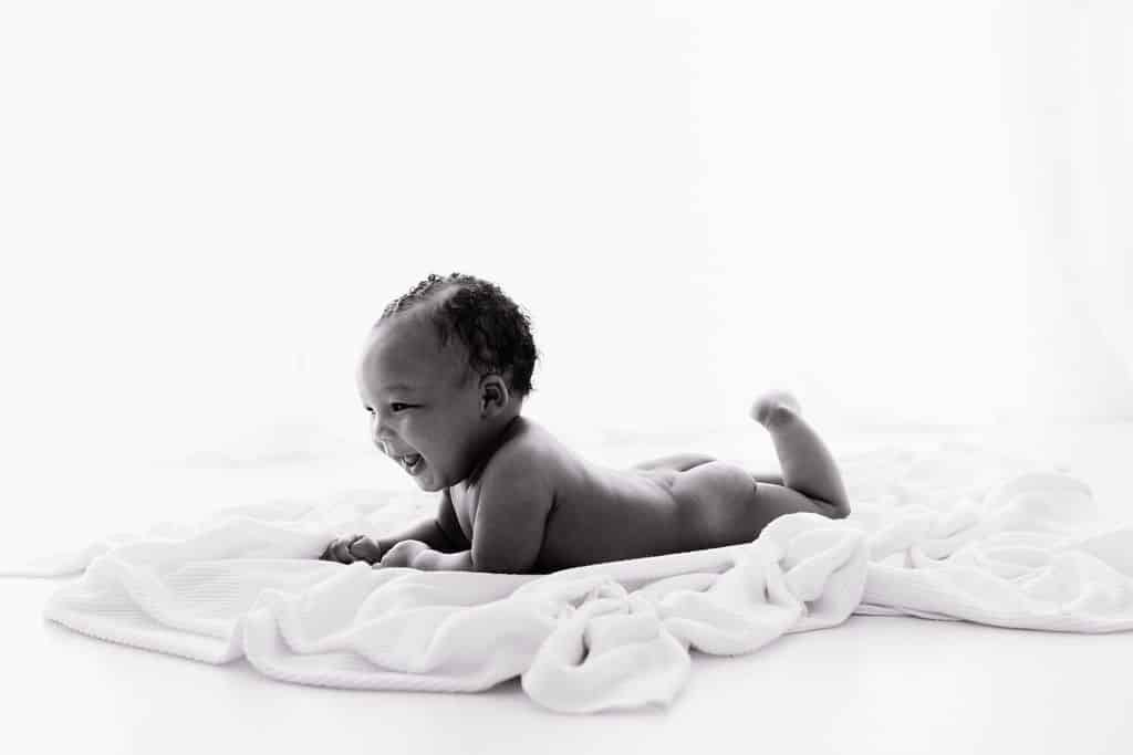 A baby lies on their stomach on a soft, white blanket, smiling and looking to the side in a bright, minimal setting.