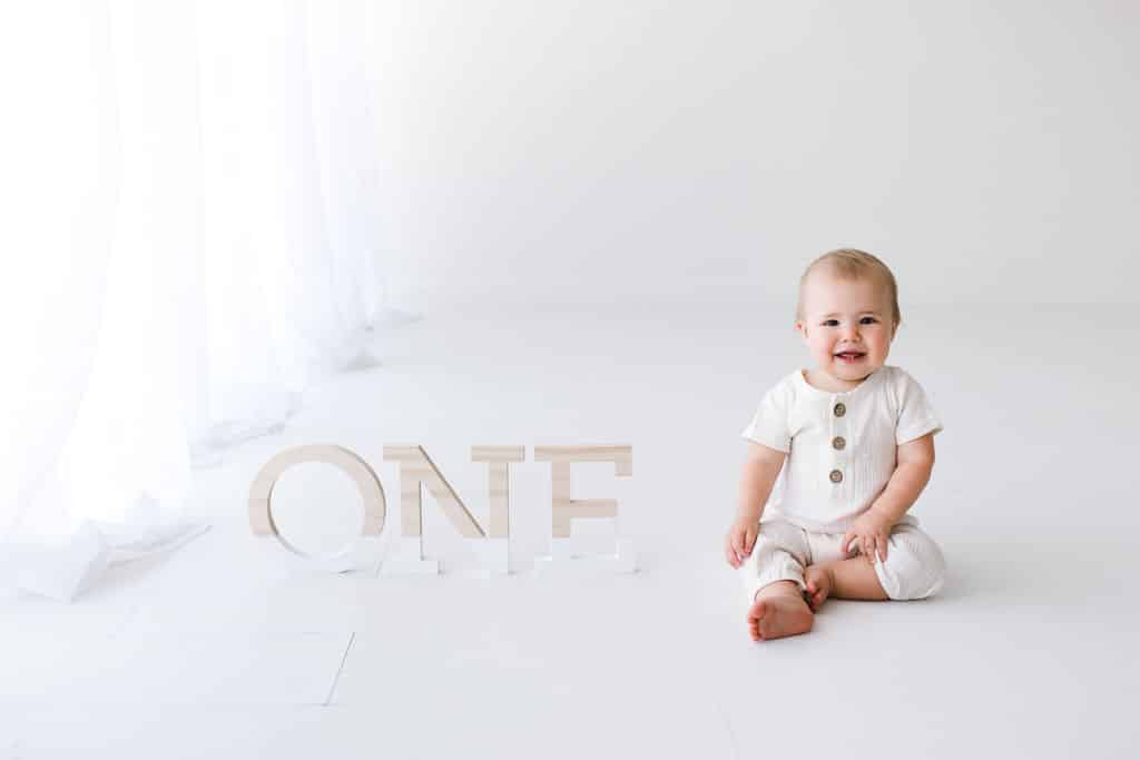A baby in a white outfit sits on the floor next to large wooden letters spelling “ONE” in a bright, white room.