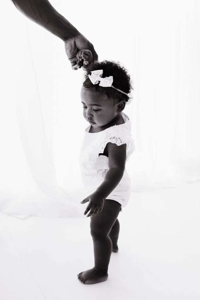 A baby in a white outfit and bow headband stands while an adult hand gently holds her hand for support.