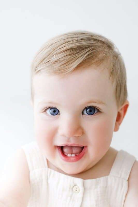 A baby with blue eyes and light hair wearing a cream-colored outfit looks at the camera with an open-mouthed smile against a white background.