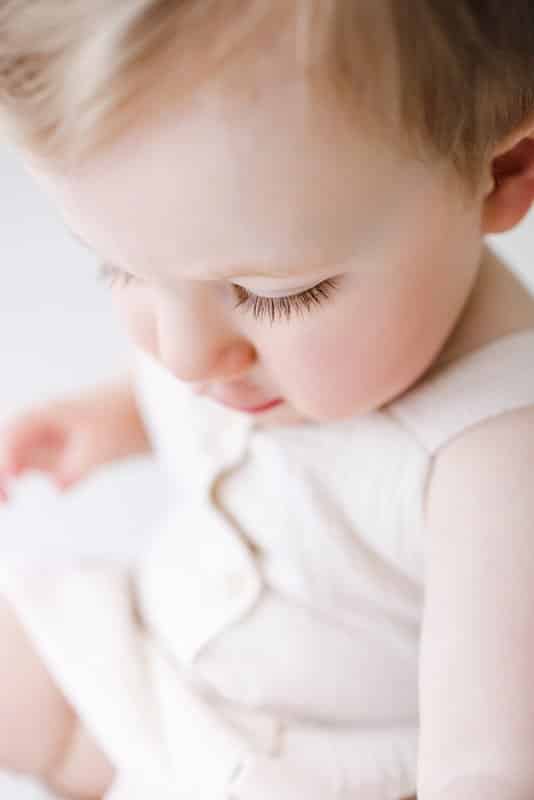 Close-up of a young child with light hair and long eyelashes, wearing a light-colored sleeveless outfit, looking down.