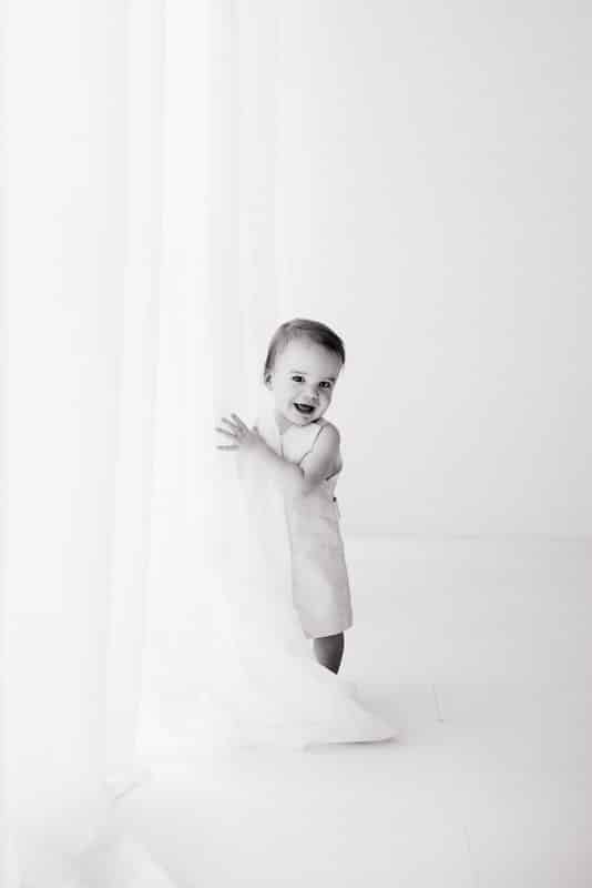 A young child stands holding a sheer curtain, smiling, in a bright, minimalist, black-and-white room.