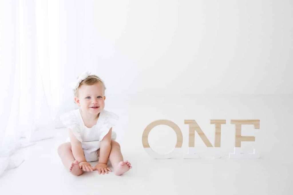 A smiling toddler in white sits on the floor next to large letters spelling “ONE” against a white background.