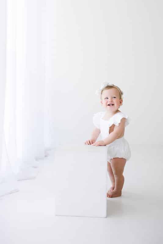 A baby wearing a white outfit stands next to a white block in a bright, minimalistic room with white curtains.
