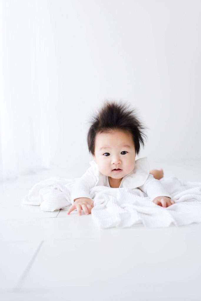 Baby with dark hair lies on a white blanket, facing forward. The background and baby’s outfit are also white, creating a soft, bright setting.