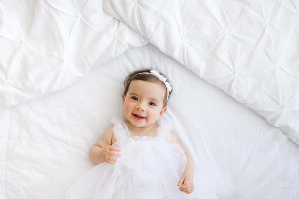 Baby wearing a white dress and headband lying on a white bed, looking up and smiling.