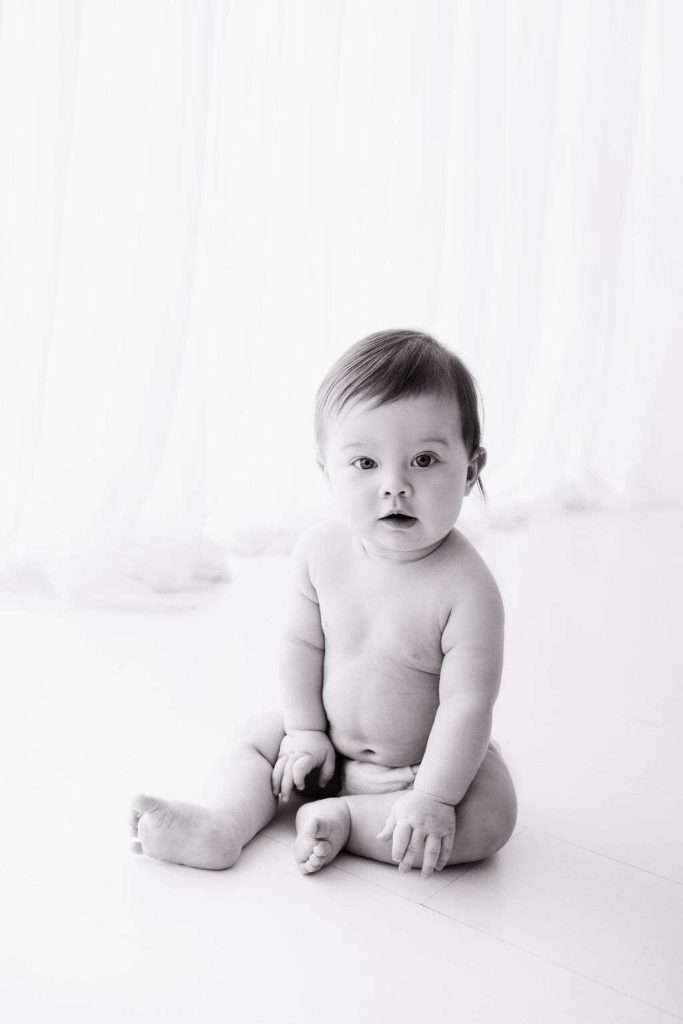 A baby with short hair sits on the floor unclothed, looking at the camera. The background is bright with sheer curtains.