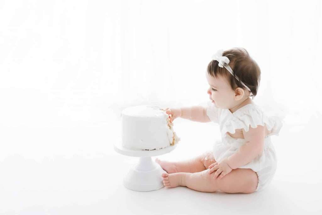 Baby in white outfit sits on floor, reaching out to touch a white frosted cake on a stand against a white background.