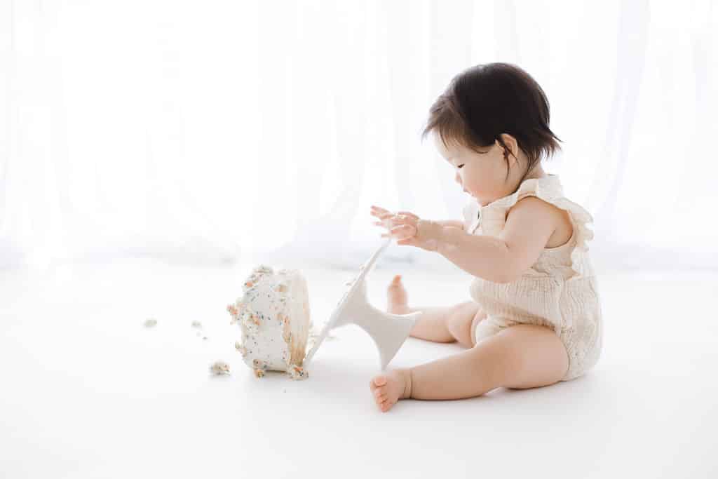 A baby in a light romper sits on a white floor, tipping over a cake with one hand. Cake and crumbs are scattered beside her.