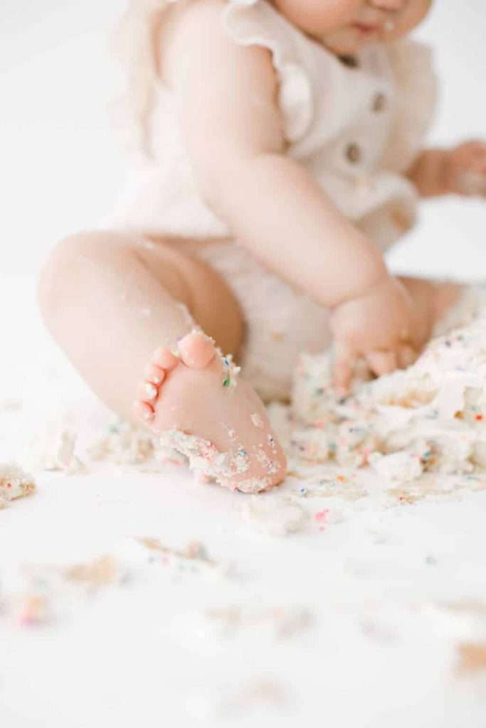 A baby in a light-colored outfit sits on the floor surrounded by smashed cake, with frosting and crumbs on their foot and hand.