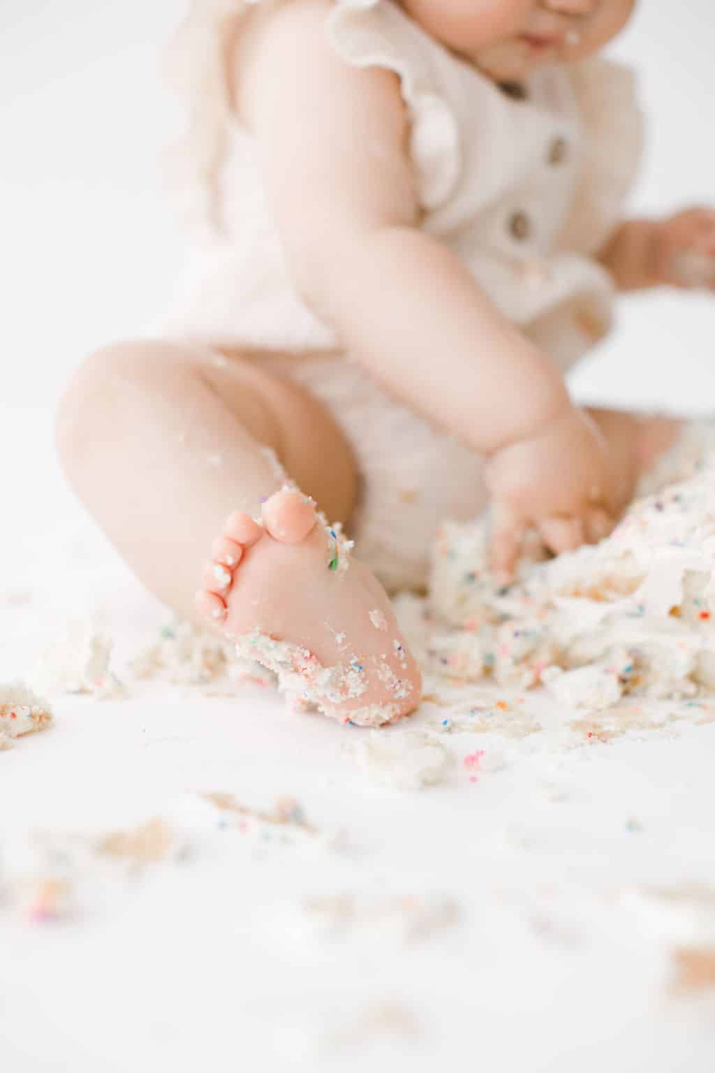 A baby in a light-colored outfit sits on the floor surrounded by smashed cake, with frosting and crumbs on their foot and hand.