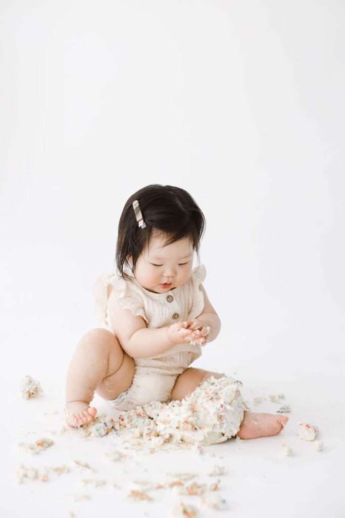 A baby sits on the floor in a white outfit, playing with and surrounded by crumbled cake on a plain white background.