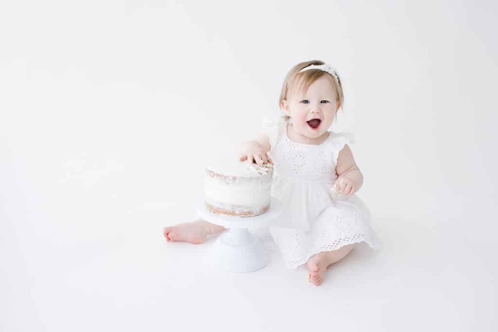 Baby in a white dress sits on the floor, touching a white frosted cake on a stand, with a plain white background.