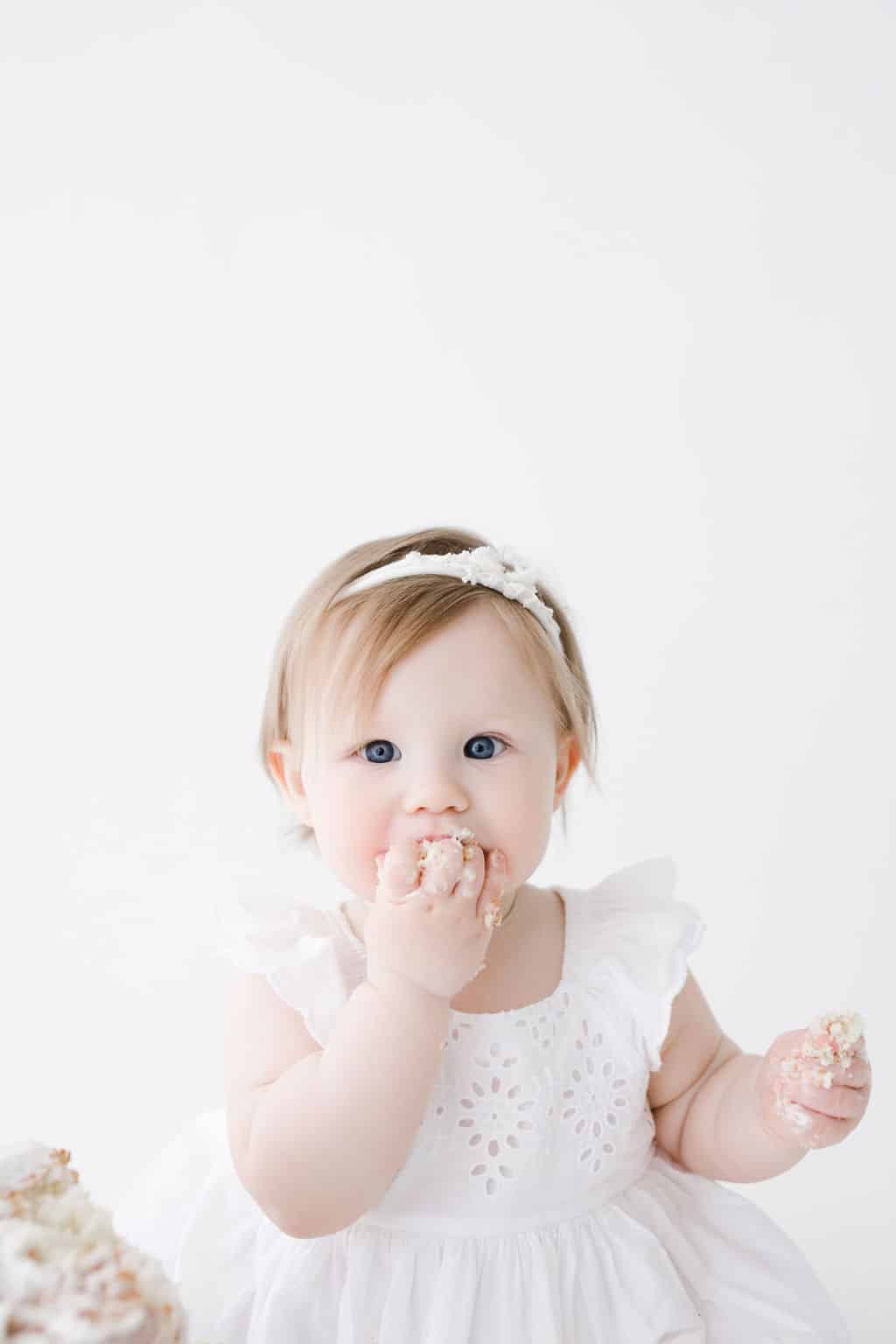 Baby in a white dress eats cake with her hands, some frosting on her face, against a plain white background.