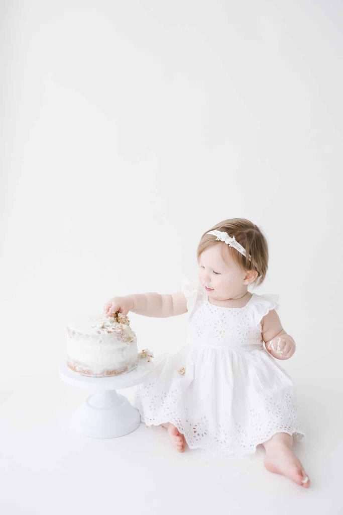 Baby in a white dress sitting on the floor, touching a white frosted cake on a stand against a plain white background.