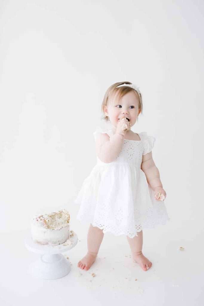 A toddler in a white dress stands barefoot on a white floor, eating cake with one hand. A partially eaten cake sits on a stand nearby, with crumbs scattered around.